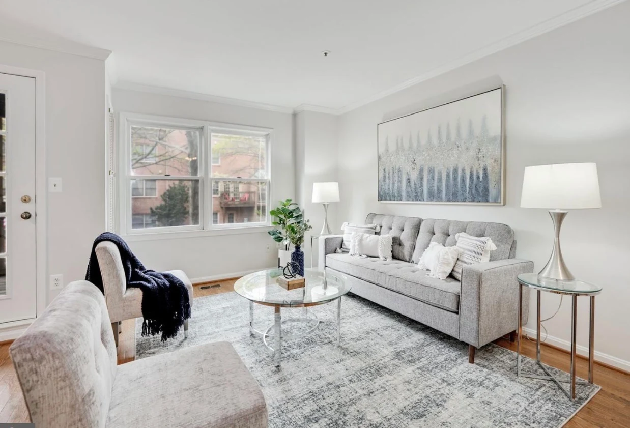 Staged living room with a tufted grey sofa, glass coffee table, and layered neutral textiles